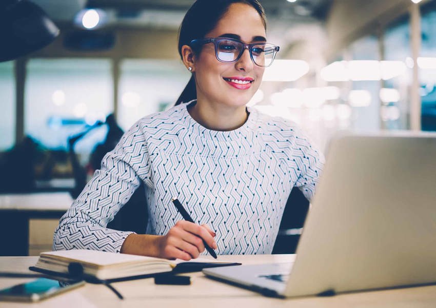 A lady in glasses sits at a desk reading something on her laptop