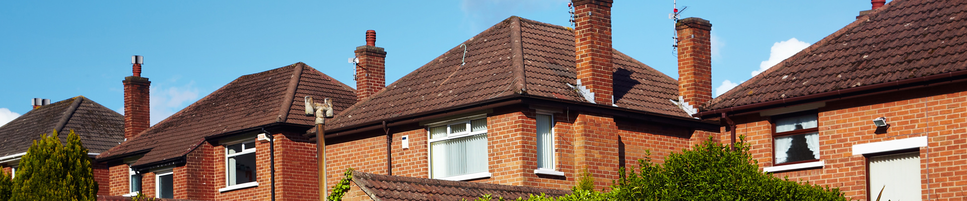 Semi-detached houses in the UK appear over a hedge, with a cloudy blue sky in the background.