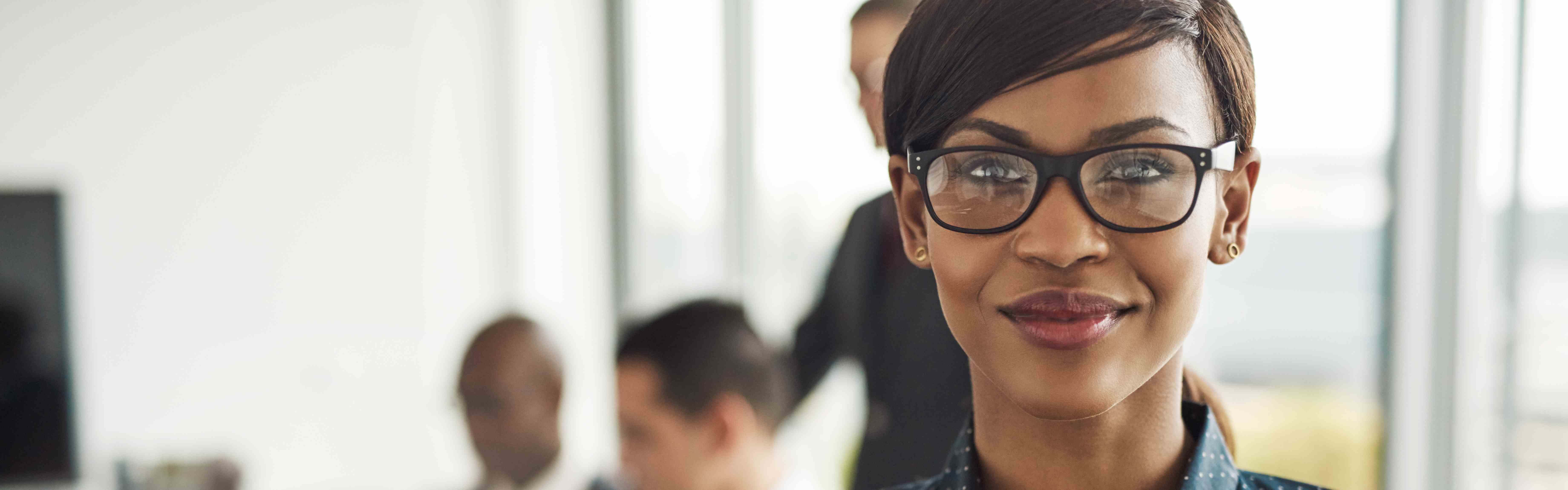 Beautiful young grinning professional Black woman in office with eyeglasses, folded arms and confident expression as other workers hold a meeting in background