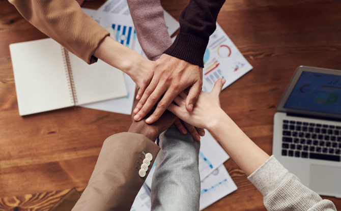 Decorative stock image of six people putting their hands in for a group celebration over a desk